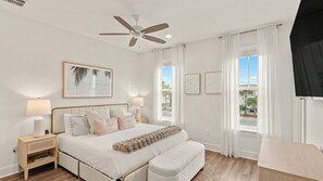 Ein Schlafzimmer mit einem Bett, Nachttischlampen, einem Deckenventilator, einem Fenster mit Vorhängen und einem gerahmten Bild an der Wand.