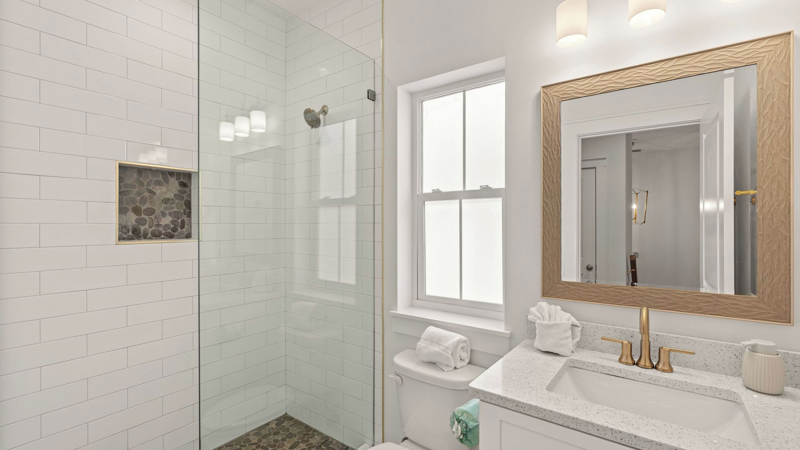 Un baño moderno con cabina de ducha de vidrio, un espejo grande con marco de madera y pared con azulejos blancos.
