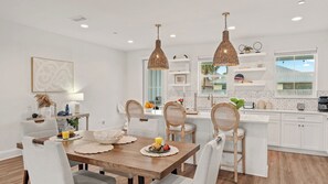 A modern kitchen with white cabinetry, a wooden dining table, wicker pendant lights, and a view of the outdoors through a window. - Low Tide by Avantstay Walk to Beach Shared Pool + Hot Tub Near St. Andrews (Panama City Beach)