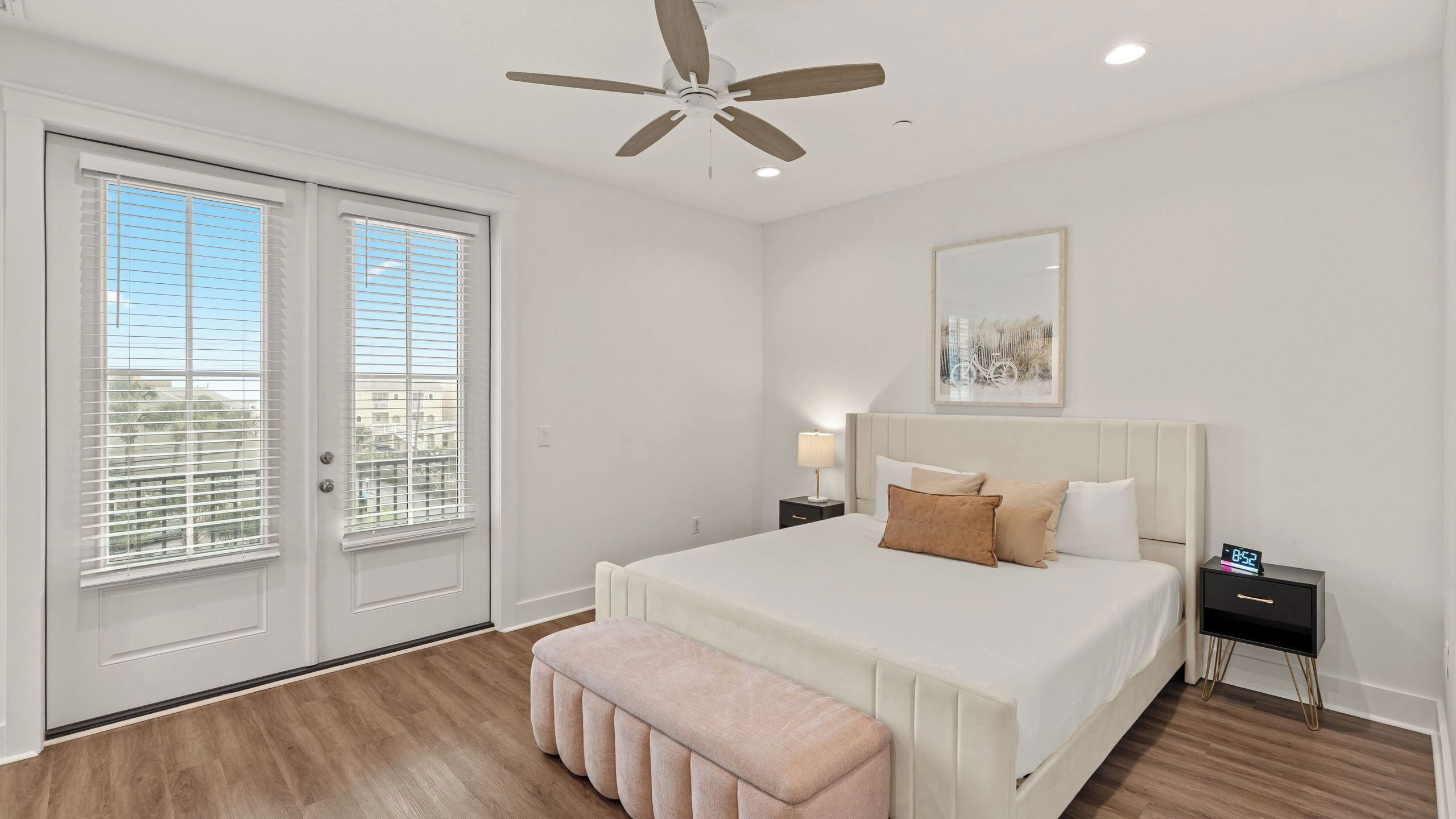 A bedroom with a bed, a bench, a nightstand with a clock, a ceiling fan, and a window with blinds.