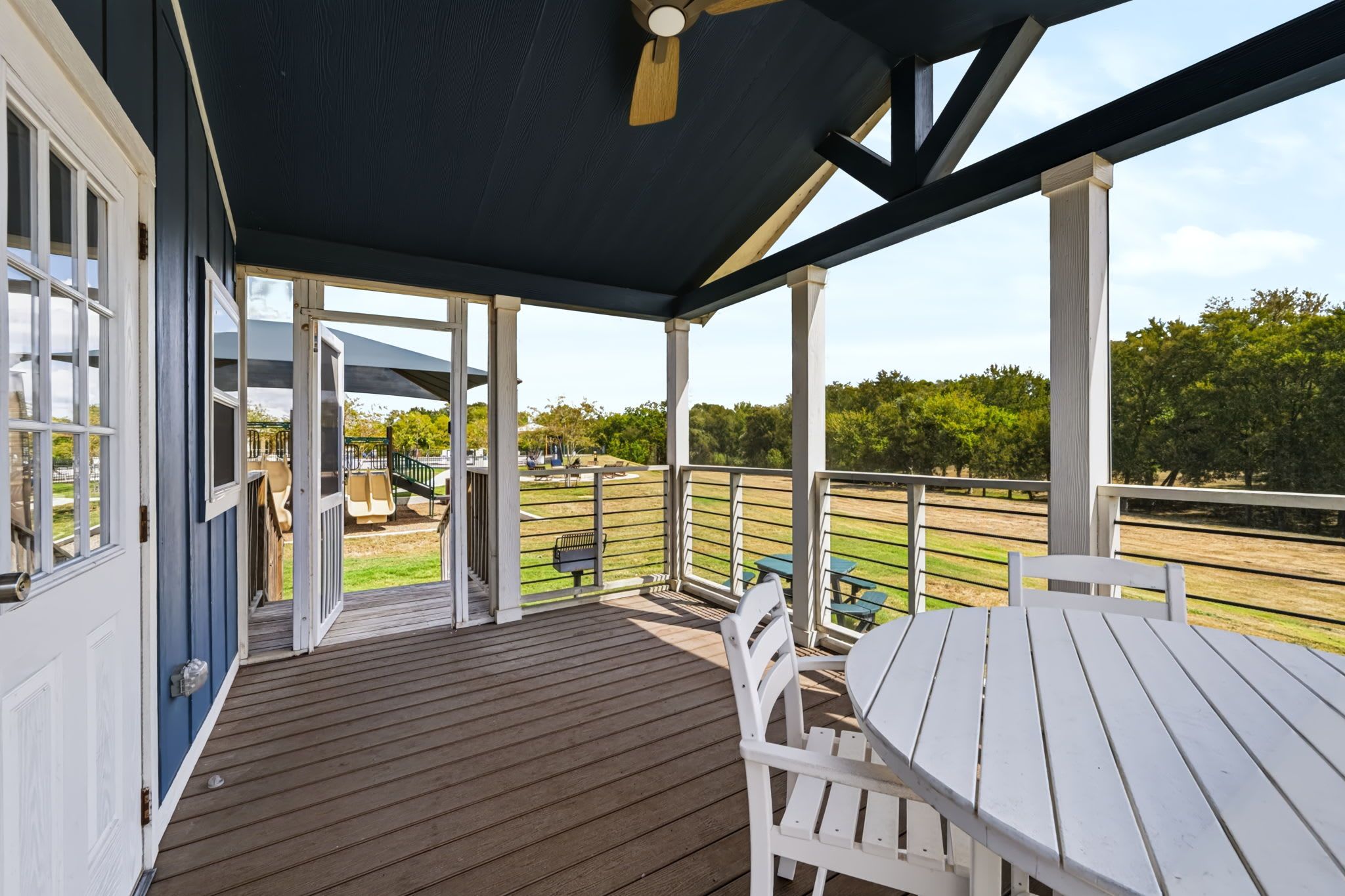 A covered outdoor area with a wooden deck, white furniture, and a view of a park with playground equipment.