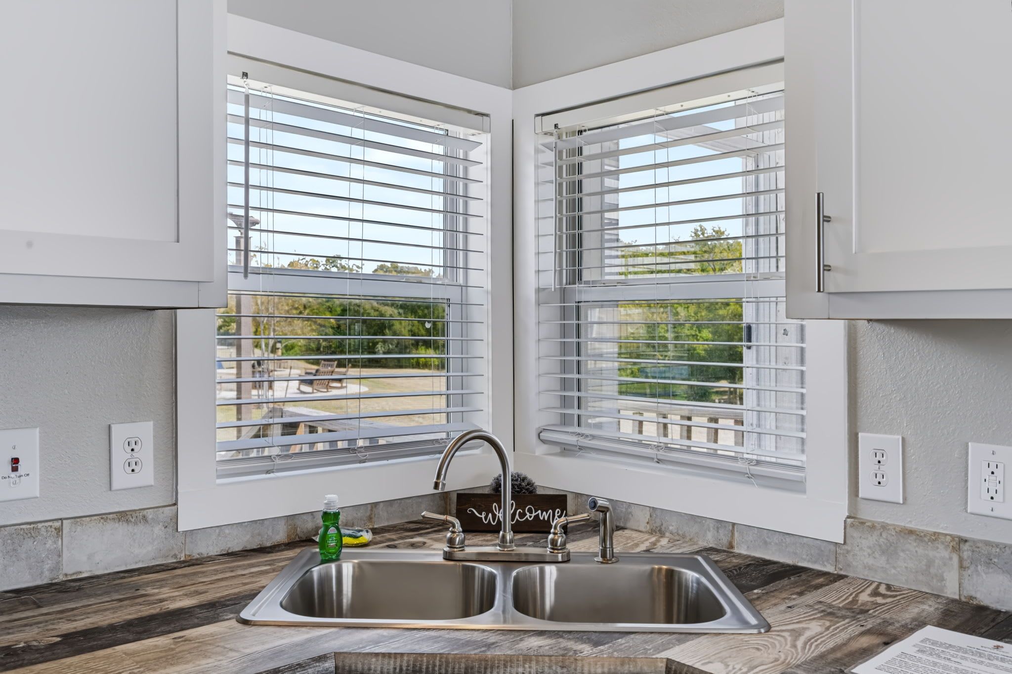 A kitchen with a double sink, two windows with blinds, and a welcome sign.