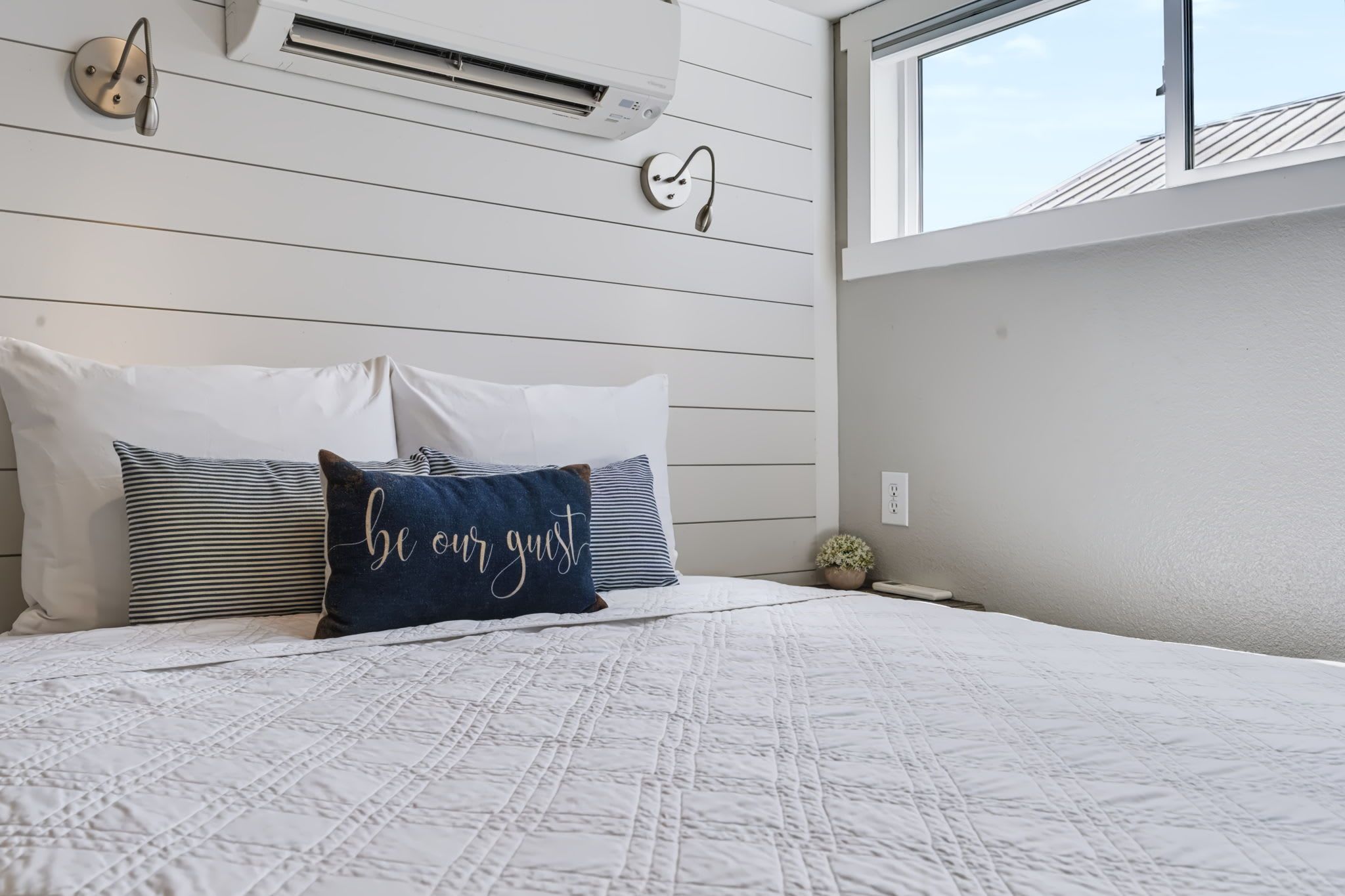 A bedroom with a bed, pillows, a wall-mounted air conditioner, a window, and a small potted plant.