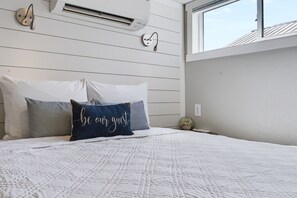 A bedroom with a bed, pillows, a wall-mounted air conditioner, a window, and a small potted plant.