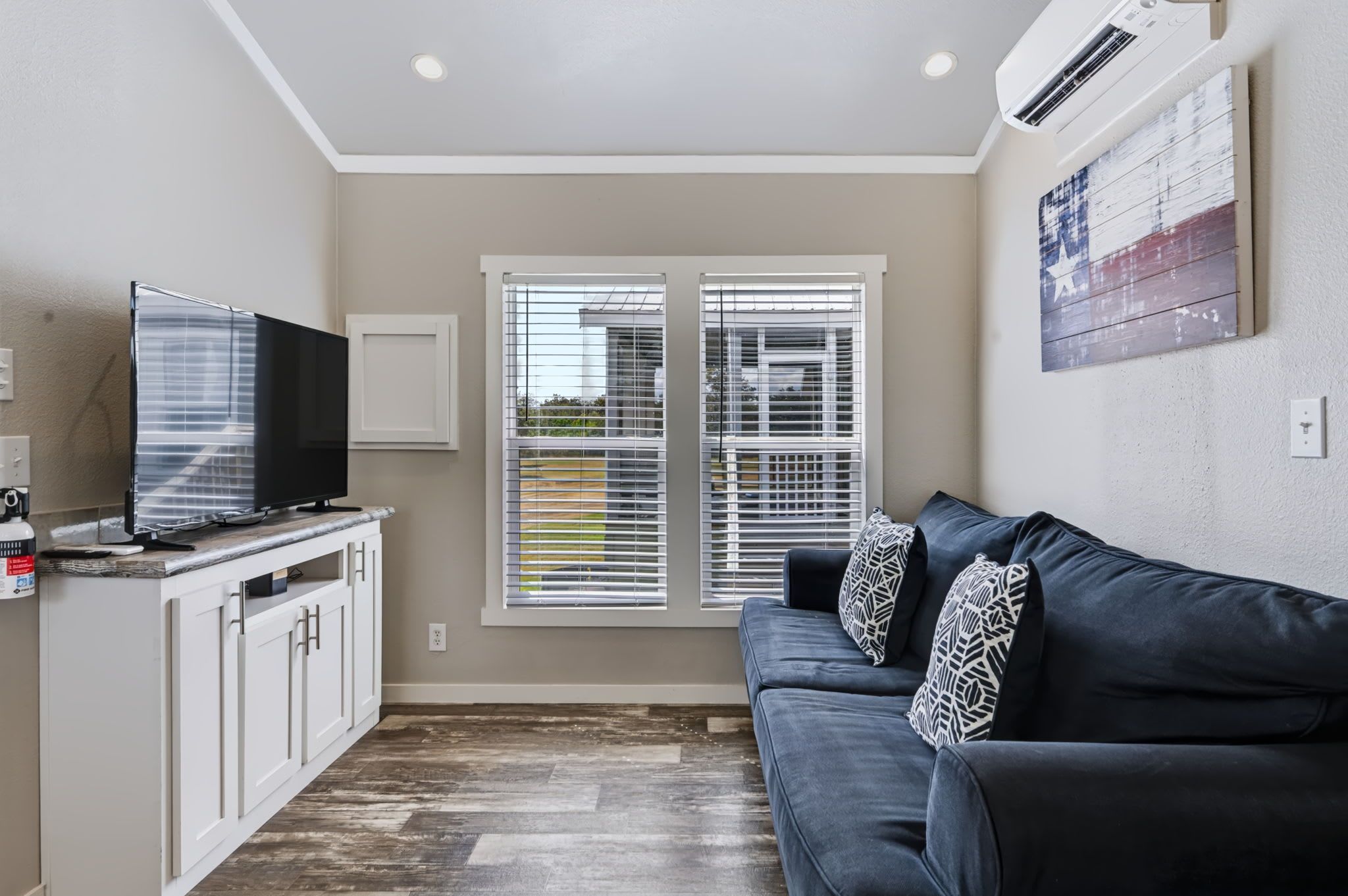 A living room with a dark sofa, a TV on a cabinet, and a window with blinds.