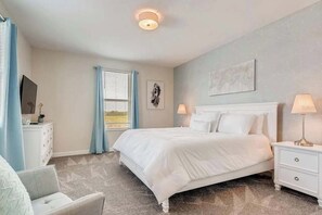 Ein Schlafzimmer mit einem Bett, einem Nachttisch, einer Kommode, einem Sessel, einem Fenster mit Vorhängen und einer Deckenleuchte.