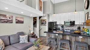 A modern kitchen with white cabinets, a central island, and a dining area with a gray sofa and wooden stools. - Serenity by the Slopes Steps to Trails + Free Bus to Slopes Balcony + Views (Vail)
