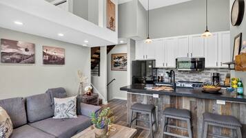 A modern kitchen with white cabinets, a central island, and a dining area with a gray sofa and wooden stools.