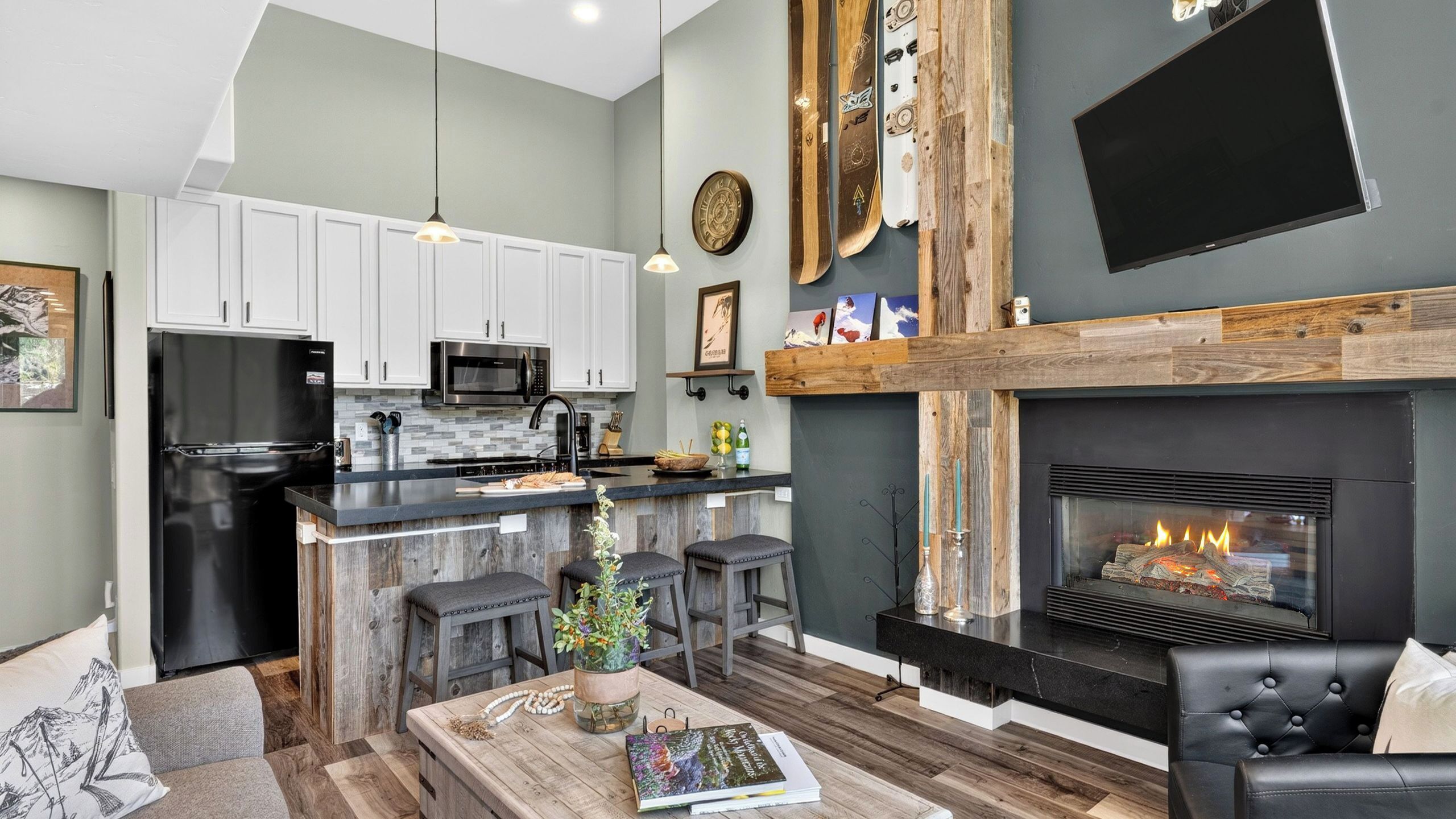 A modern kitchen with white cabinets, a black refrigerator, and a microwave. There's a wooden island with stools, a fireplace, and a flat-screen TV mounted on the wall.