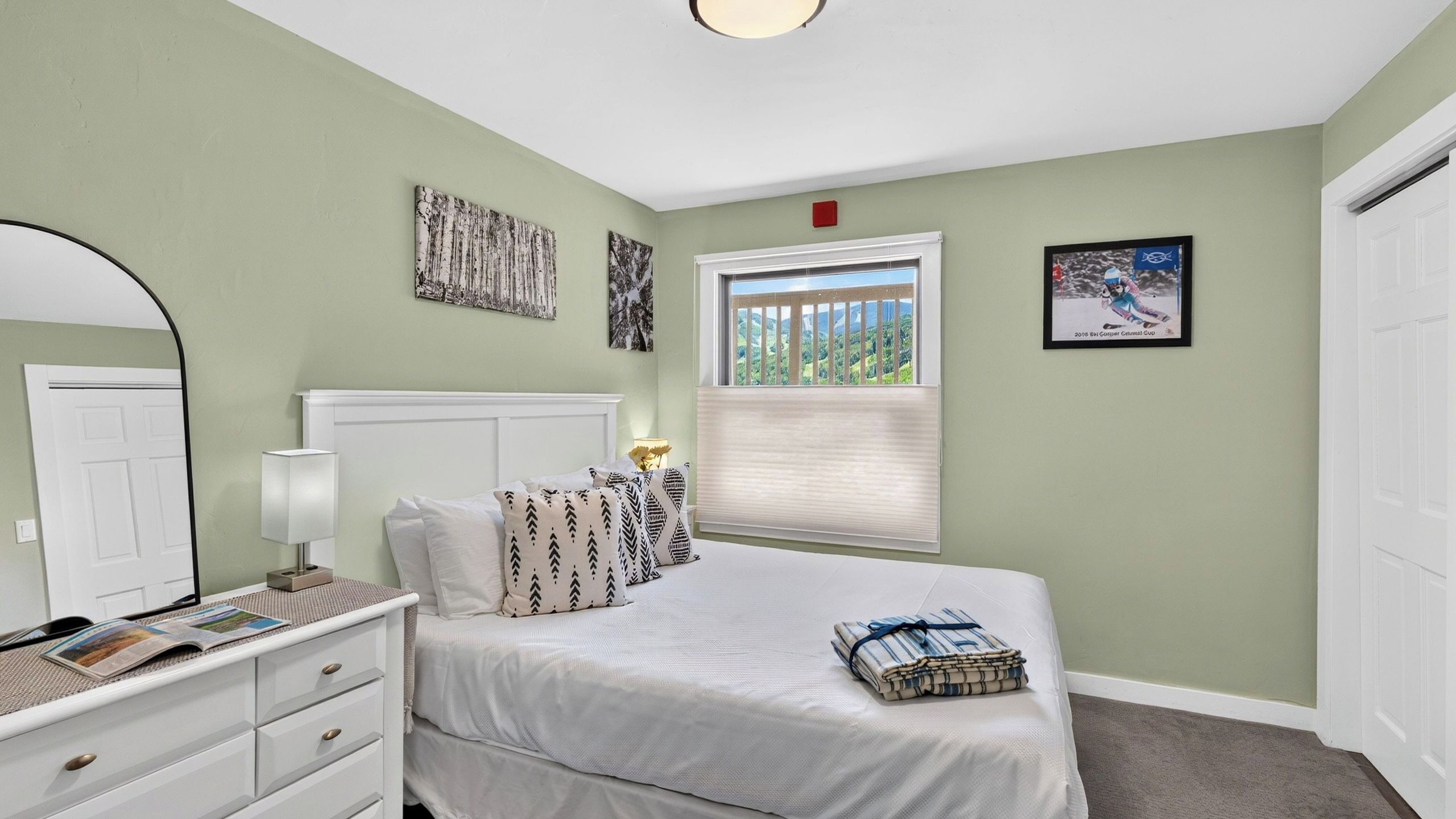 A bedroom with a bed, dresser, mirror, window with blinds, and artwork on the walls.