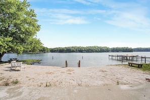 A lakeside beach with a wooden dock extending into the water, a picnic table, and a bench.