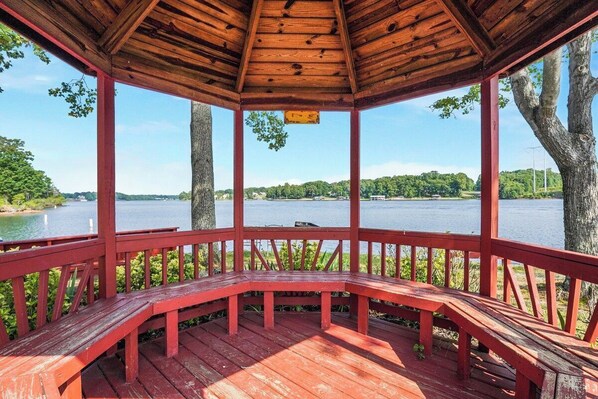 A wooden gazebo with a bench, overlooking a lake with trees and a clear sky.