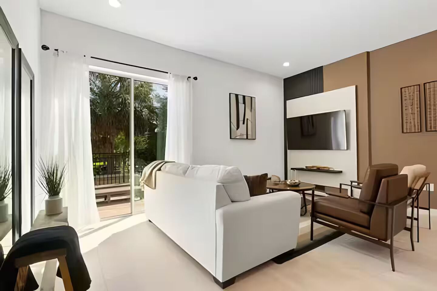 A modern living room with a sofa, a dining table, and chairs. There is a sliding glass door leading to a balcony.