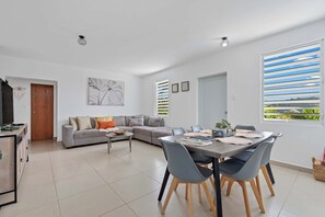 A modern living room with a grey sofa, a dining table with chairs, and a large window with blinds. - Casa Brisa w/ Heated Pool + Mt. View + River (Las Marías)