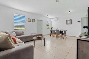 A modern living room with a grey sofa, a dining table with chairs, and a kitchen area with a TV and remote controls.