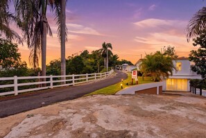 A residential street with a white fence, palm trees, and houses with lit windows.
