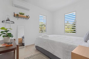 A bedroom with a bed, a bedside table, a chair, a window, and a wall-mounted air conditioner. - Casa Brisa w/ Heated Pool + Mt. View + River (Las Marías)