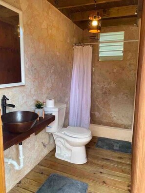 A compact bathroom with a wooden floor, a white toilet, a wooden basin, a hanging light fixture, and a window with blinds.