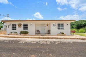 A single-story building with a flat roof, a front porch with two chairs, and a small garden area with potted plants. - Casa Verde at Hacienda Serena w/ Mt. View + River (Las Marías)