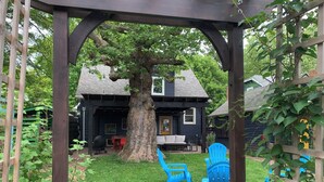 Un espacio de estar al aire libre cubierto con una pérgola, un árbol y una casa al fondo.