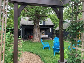 Ein überdachter Außenbereich mit Pergola, einem Baum und einem Haus im Hintergrund.