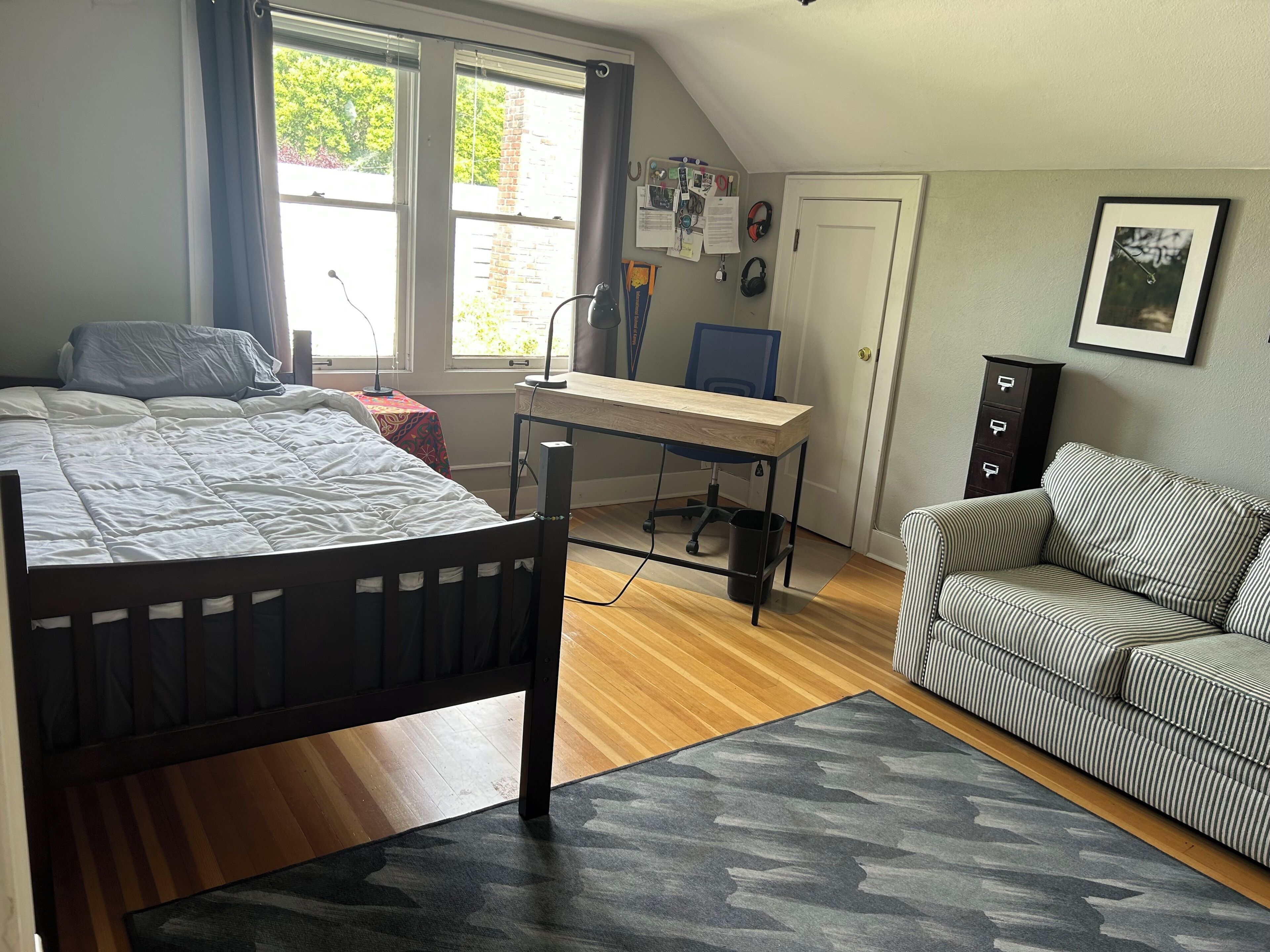 A bedroom with a bed, a desk, a chair, a sofa, and a window with curtains.