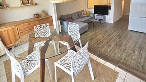 Un salon comprenant une table à manger en verre, des chaises blanches, un canapé, un téléviseur et un coin cuisine avec un réfrigérateur.