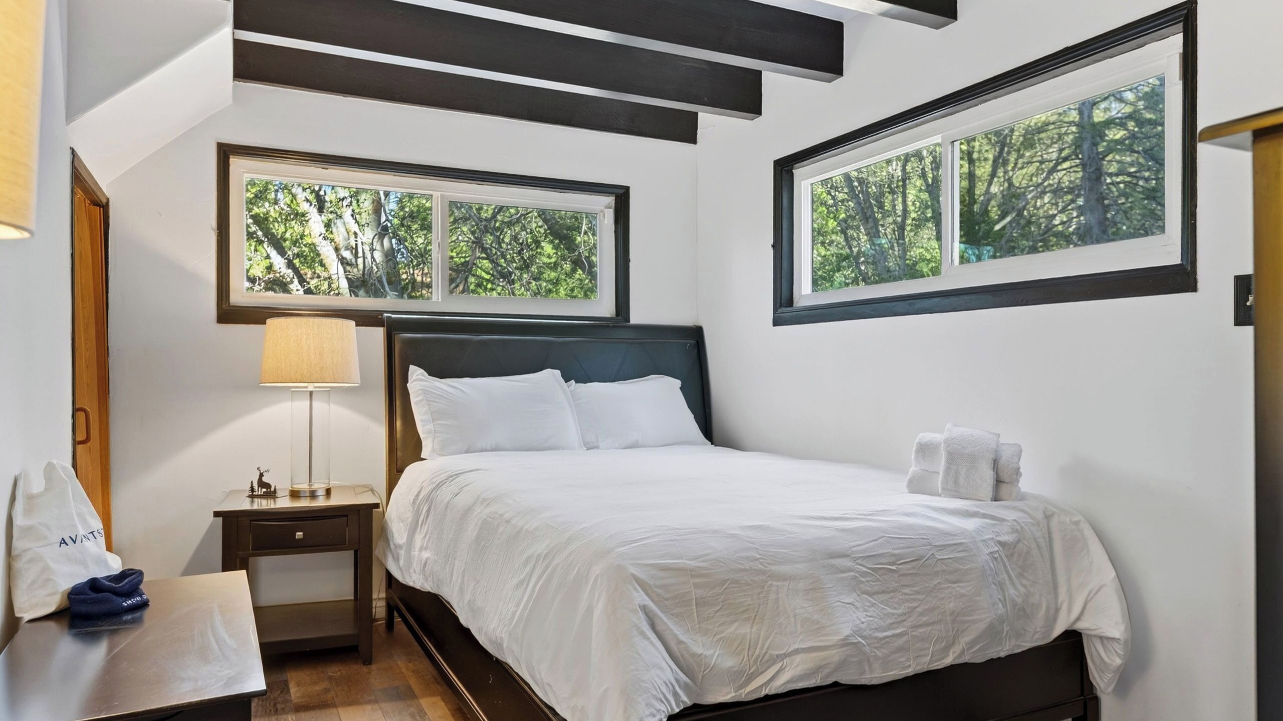 A bedroom with a bed, two bedside tables, a nightstand, a lamp, and two windows with a view of trees.
