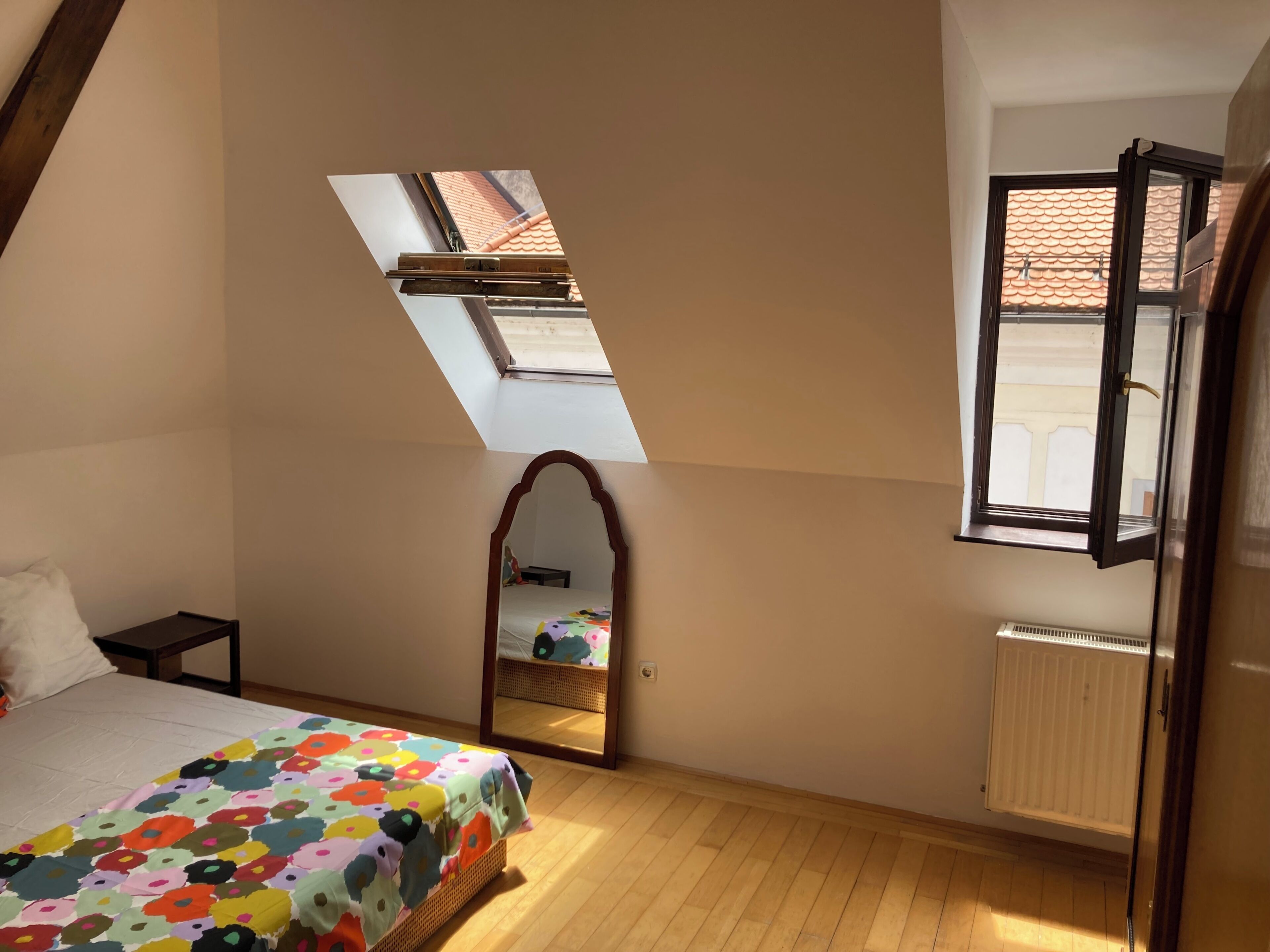 Une chambre avec un plafond en pente, un puits de lumière, un lit recouvert d’une courtepointe à motifs floraux colorés, un miroir et un radiateur.
