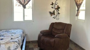 Una habitación con una cama, un sillón de cuero marrón, un adorno en la pared y dos ventanas con cortinas.