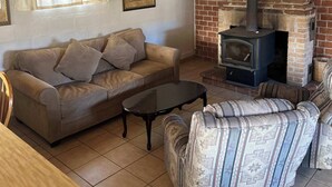 A cozy living room with a brick fireplace, wooden beams, a sofa, a patterned armchair, and a table.