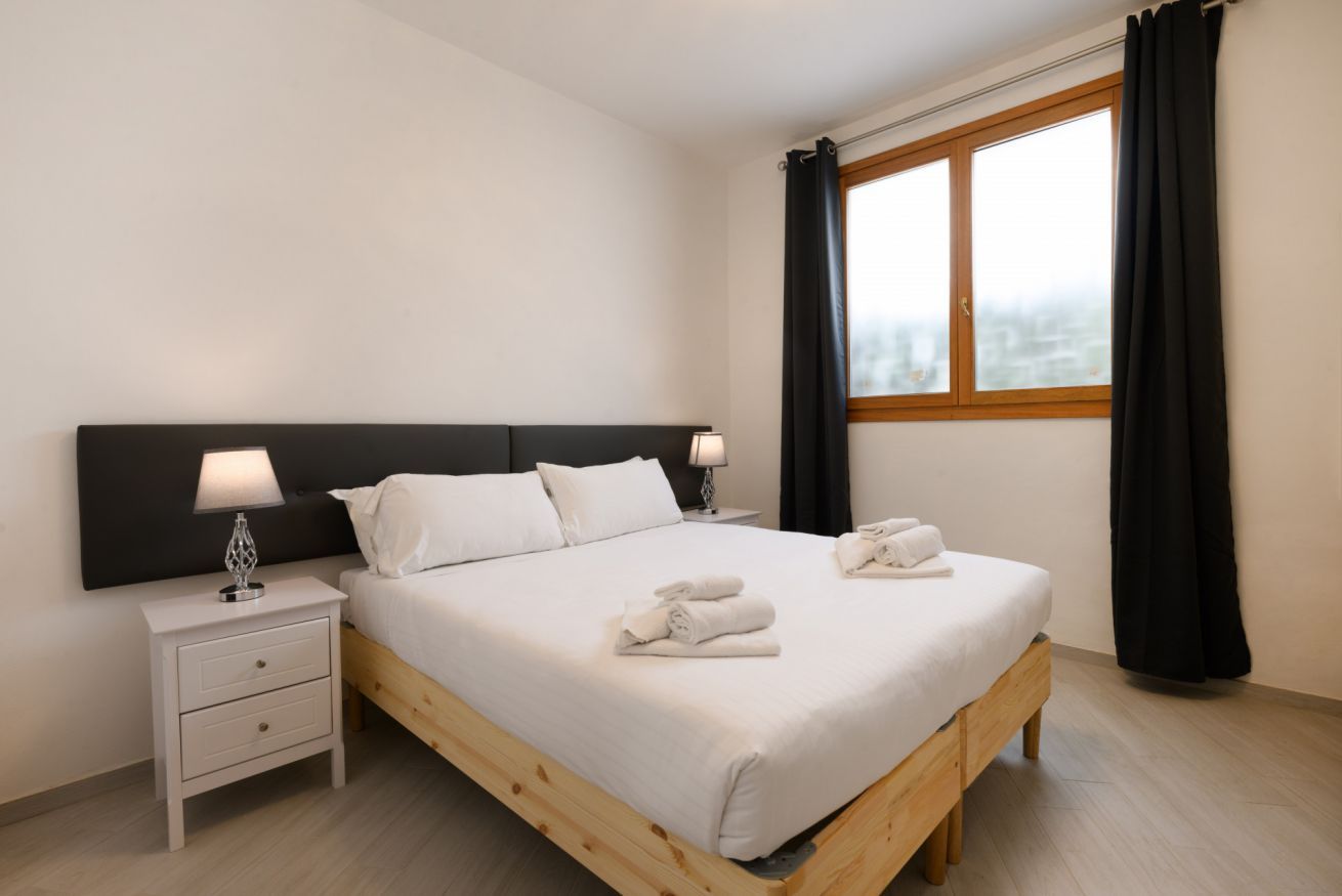 Una cama bien hecha con ropa blanca y dos toallas, una mesita de noche con lámpara y una ventana con cortinas.