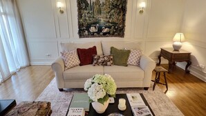 A living room with a sofa, a coffee table with books and a vase of flowers, a painting on the wall, and a lamp on a side table. - Elegant Historic Duplex in Old Town • Lincoln Park (Chicago)
