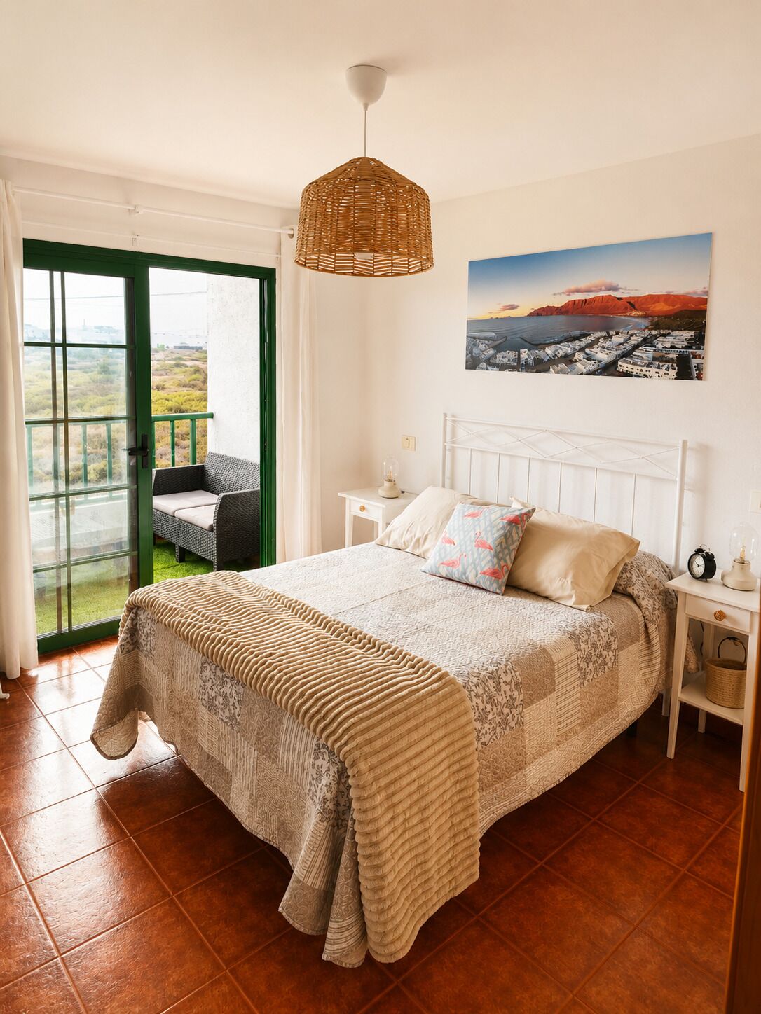 Quarto com cama, mesa de cabeceira, luminária de vime pendente, quadro e varanda com cadeira.