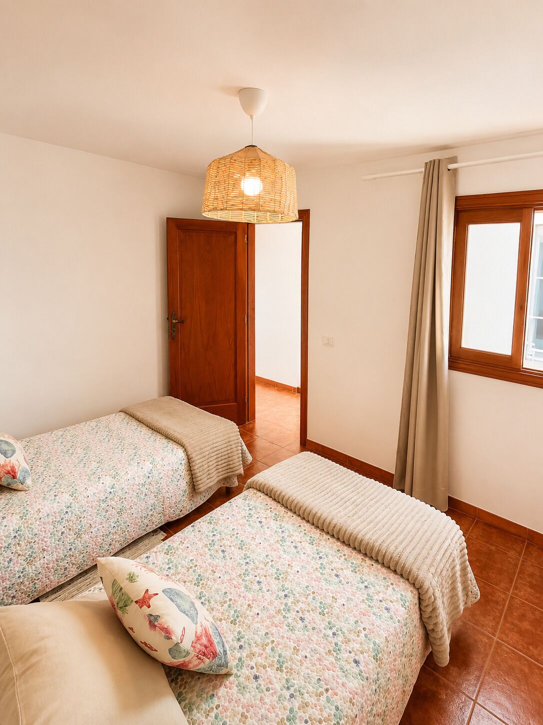 A room with two beds, a door, a window with curtains, and a hanging light fixture.
