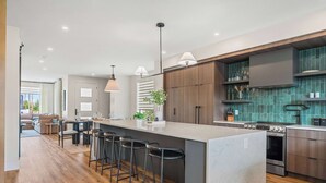 A modern kitchen with a central island, bar stools, and a dining area with a sofa and table. - The Shoreline | Luxury Escape w/ Rooftop & Hot Tub (Sylvan Lake)