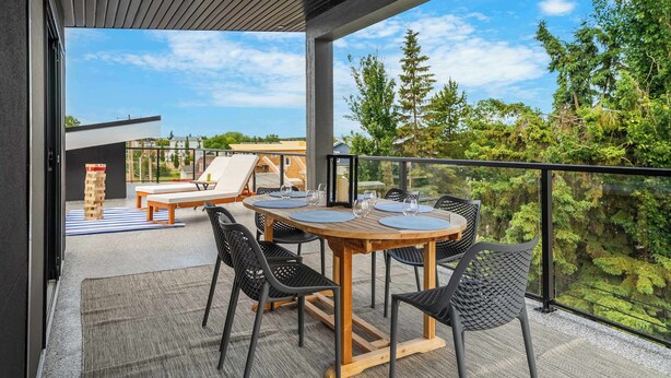 A modern outdoor patio with a wooden table and chairs, a glass balustrade, and a view of trees and buildings. - The Shoreline | Luxury Escape w/ Rooftop & Hot Tub (Sylvan Lake)