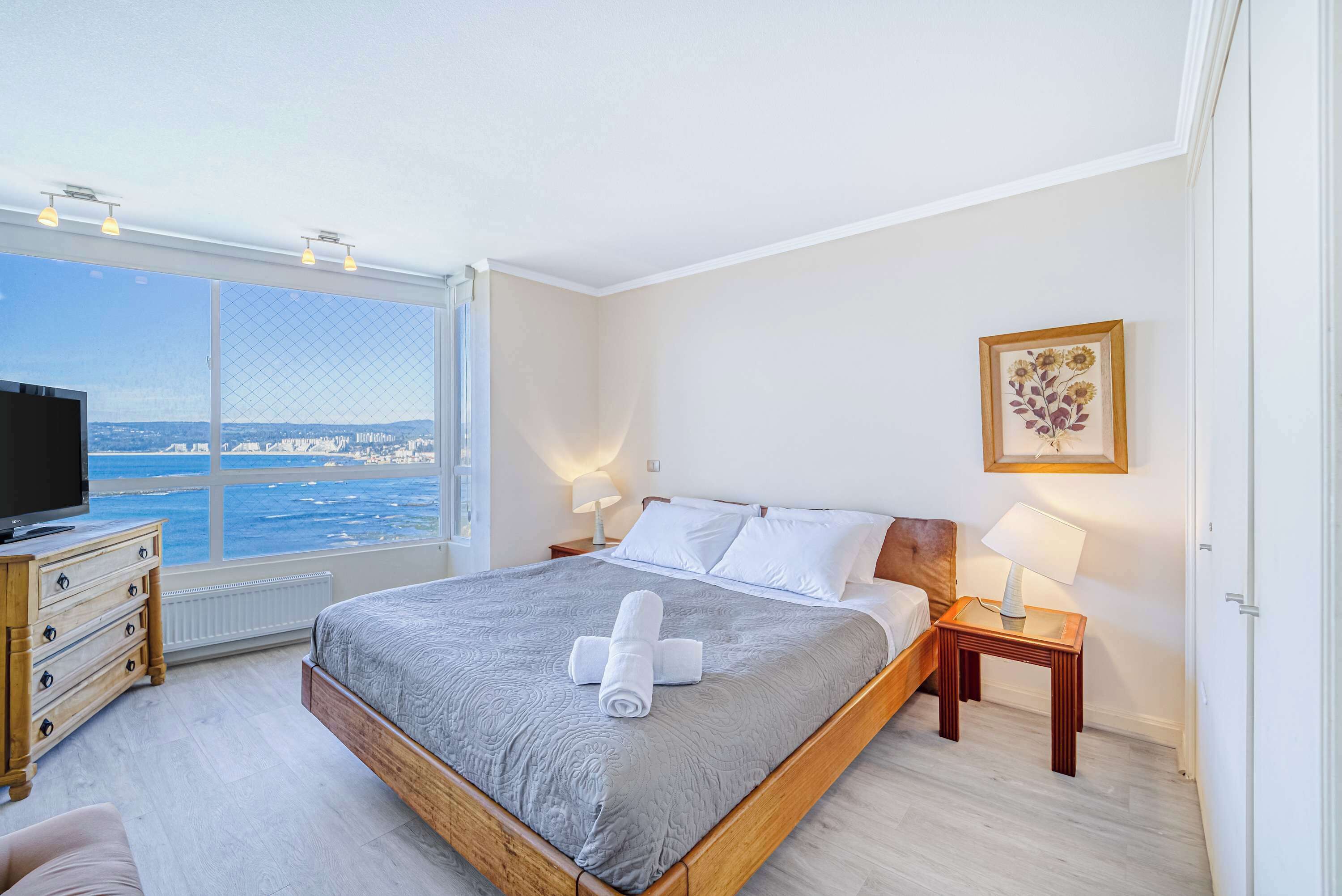 A bedroom with a large window, a bed, a dresser, a TV, and a framed picture on the wall.