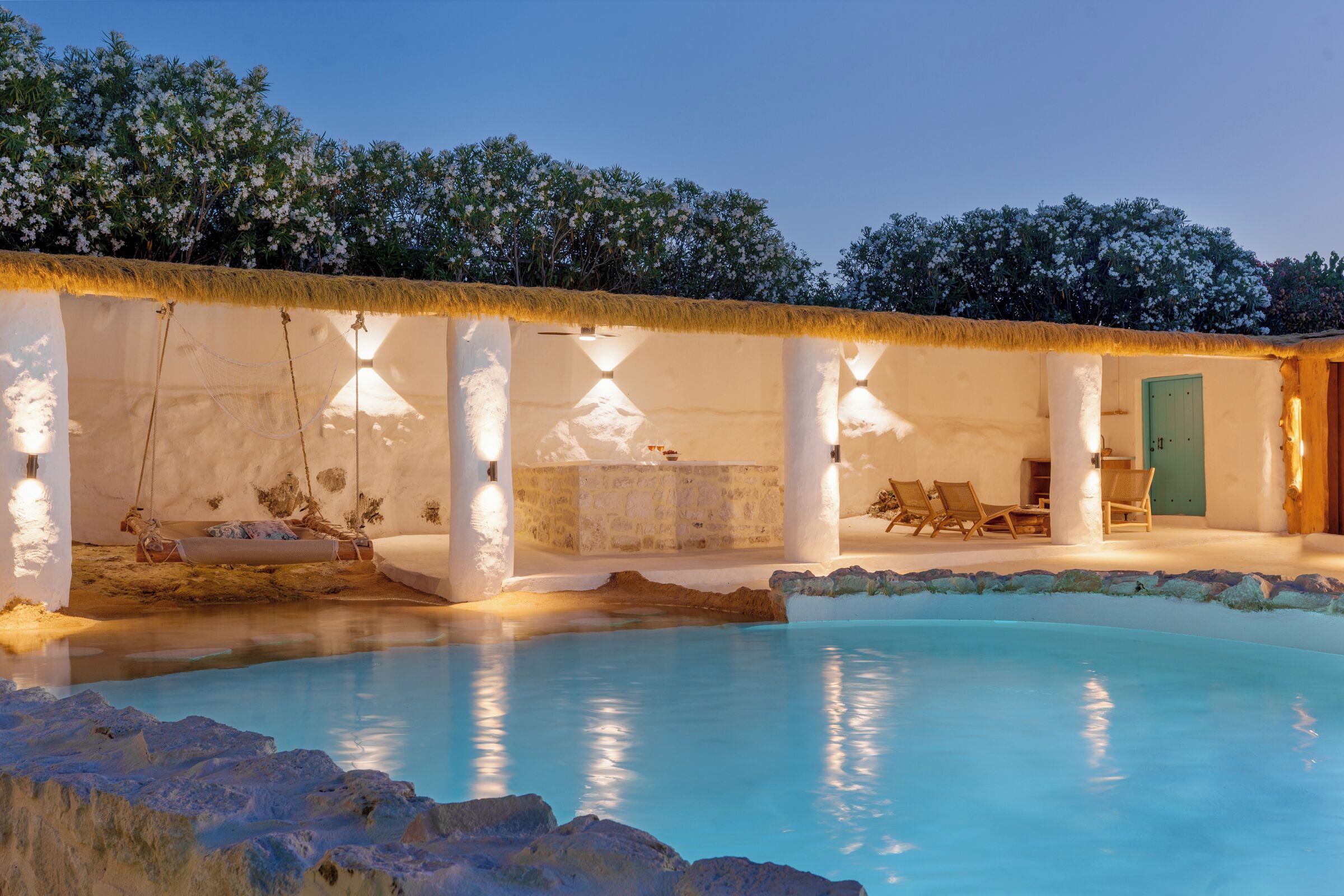 A pool with a thatched roof, a lounge area with chairs, and a stone wall.