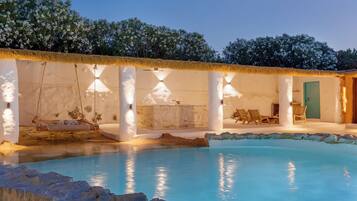 A pool with a thatched roof, a lounge area with chairs, and a stone wall.