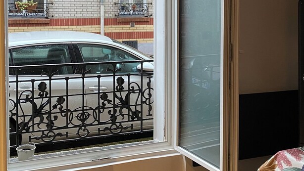 Een kamer met een groot raam, houten visgraatvloer, een zwart metalen balkonhek en een hanglamp.