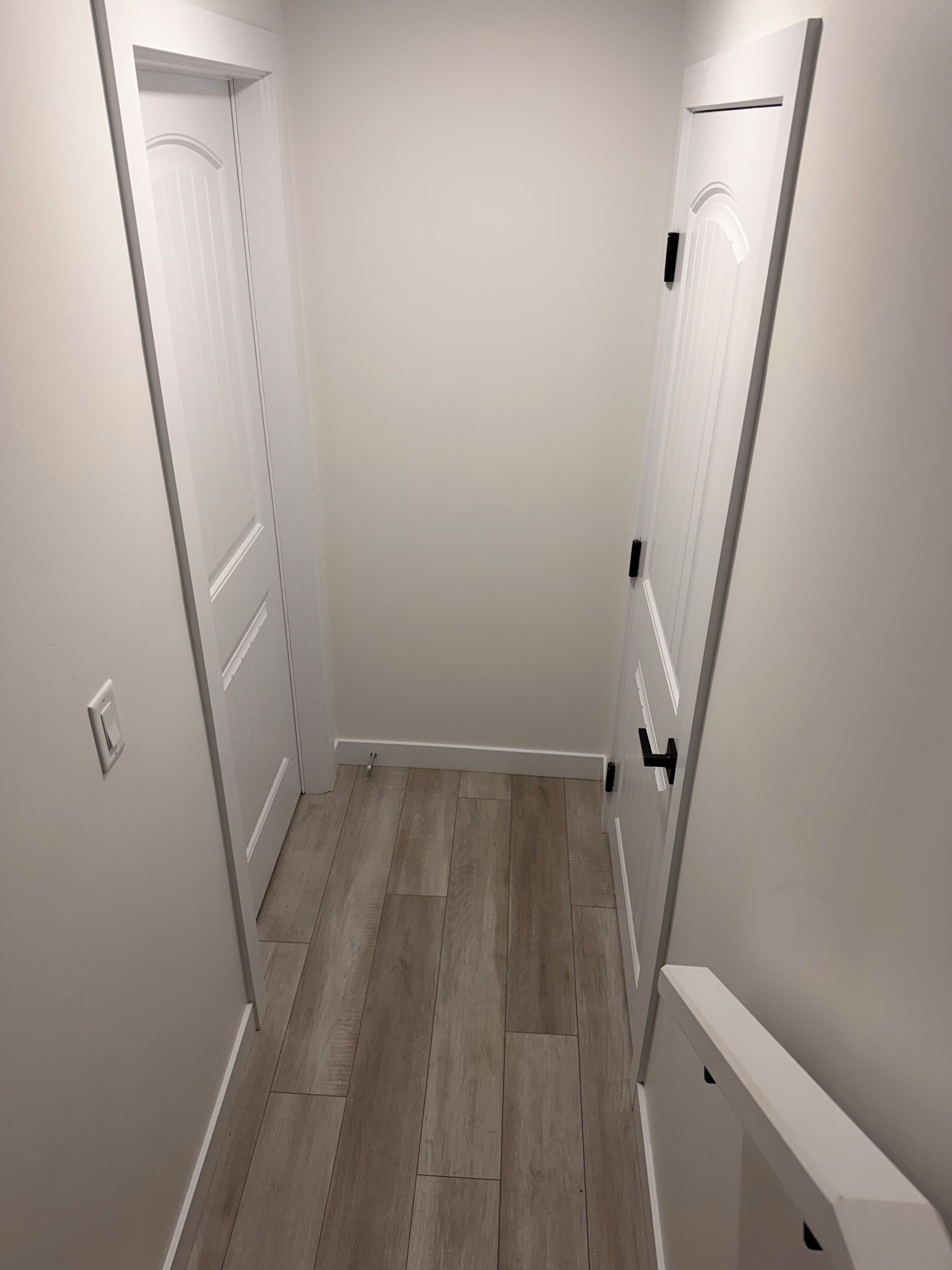 A narrow hallway with white walls and wooden flooring, featuring two white doors with black handles.