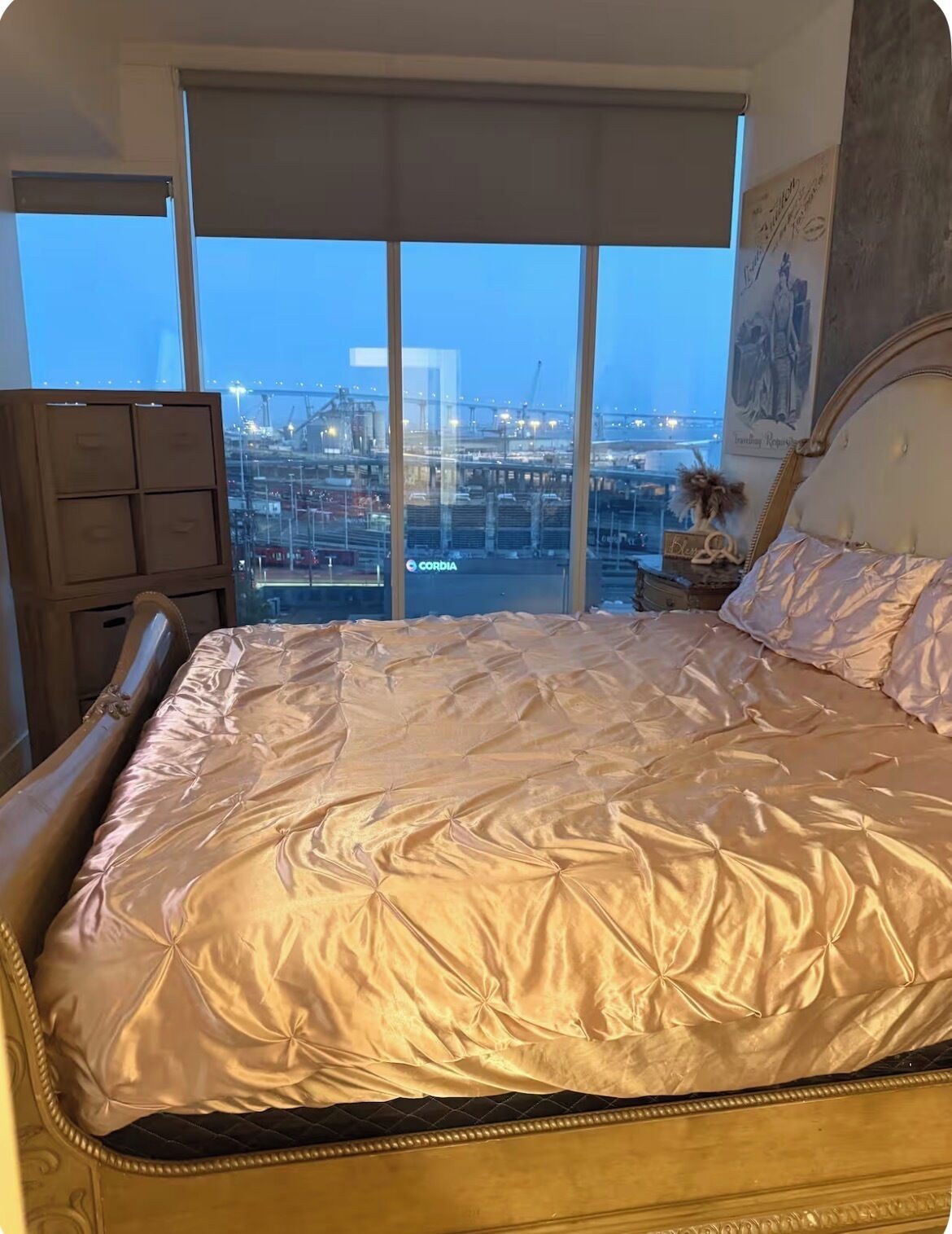 A bedroom with a large bed, a dresser, and a view of an industrial area through floor-to-ceiling windows.
