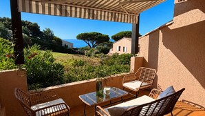 A covered outdoor seating area with wicker furniture, a glass table, and a view of the sea and greenery. - Les Coteaux de la Nartelle, air-conditioned studio with ocean view, pool, sleeps 4 (Sainte-Maxime)
