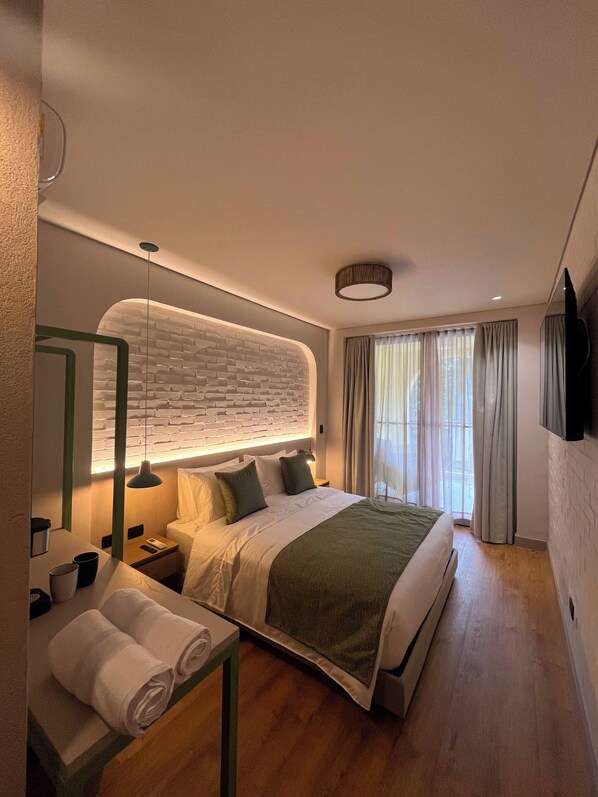 Una habitación de hotel moderna con una cama grande, una mesita de noche con lámpara, un espejo y vistas a la ciudad a través de la ventana.