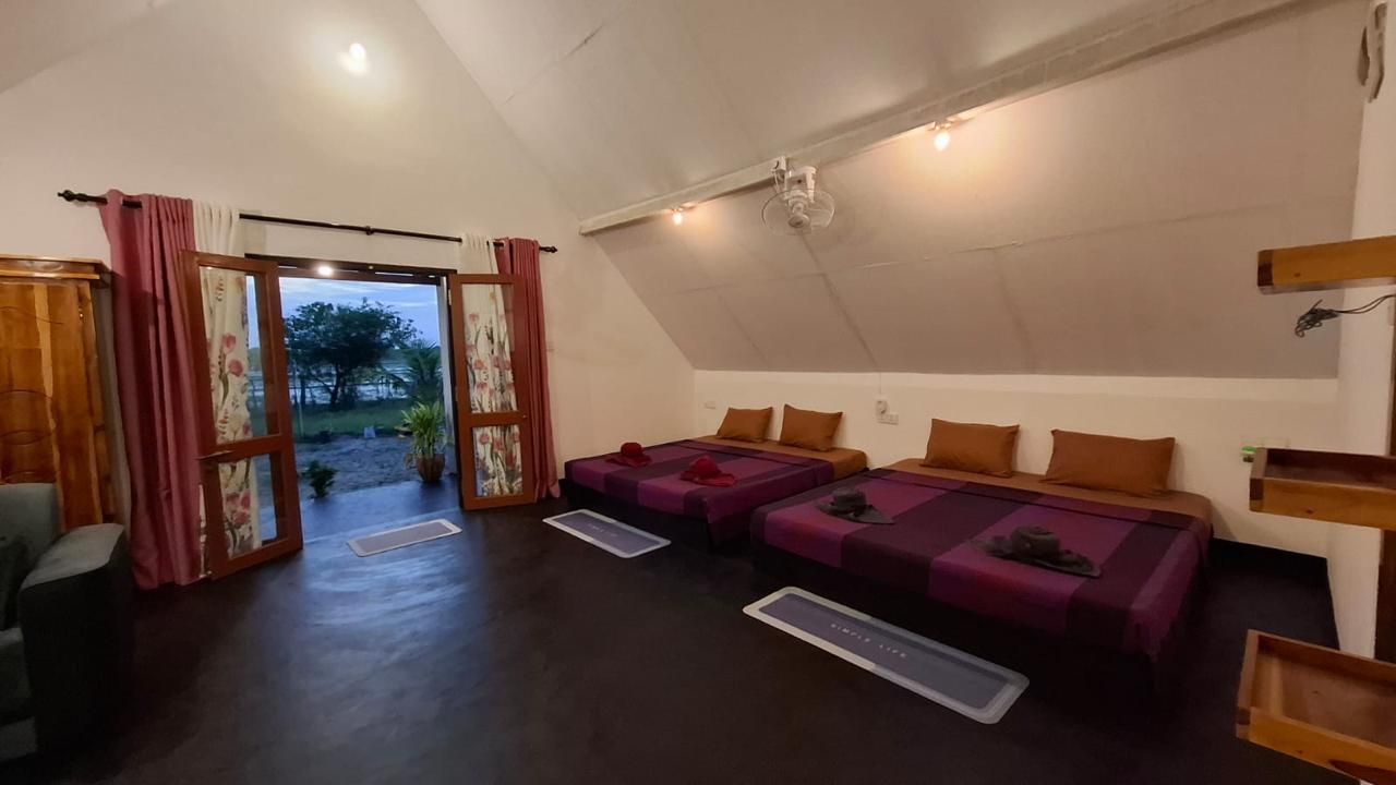 Une pièce avec un plafond en pente, deux lits avec une literie violette, une table de chevet en bois et une vue sur l’extérieur, visible à travers une porte.