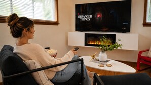 A person sitting on a couch watching TV in a room with a fireplace, a plant, and a red chair.