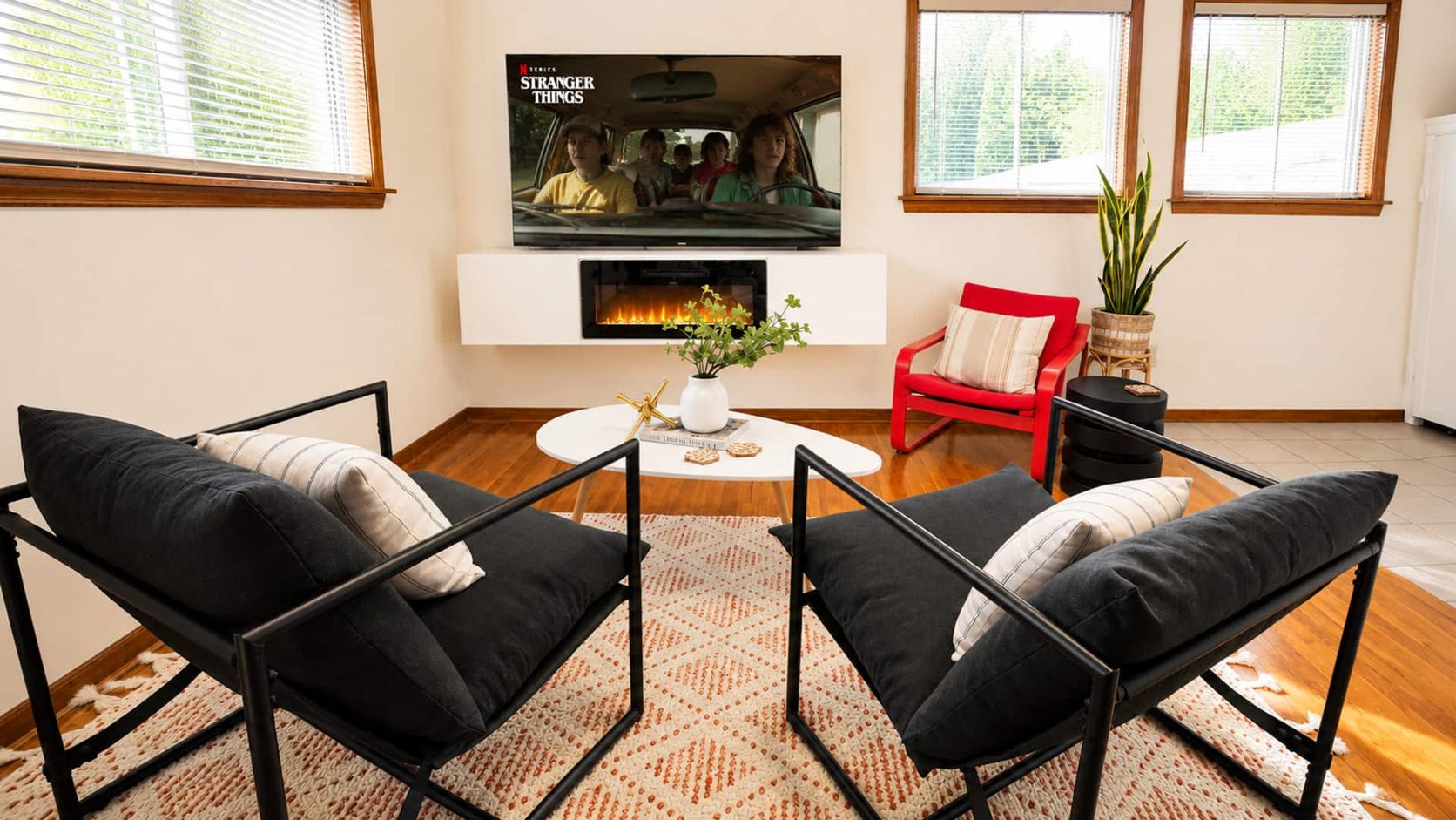 A living room with a flat-screen TV mounted above a fireplace, two black chairs with cushions, a red chair, a small table with a plant, and a window with blinds.