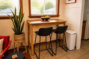 A compact kitchen with a wooden table, two black bar stools, a white trash bin, a wicker plant, and a red chair.