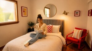 A person sitting on a bed reading a book, with a bedside table, a mirror, and a red chair in a room with a window and framed pictures on the wall.